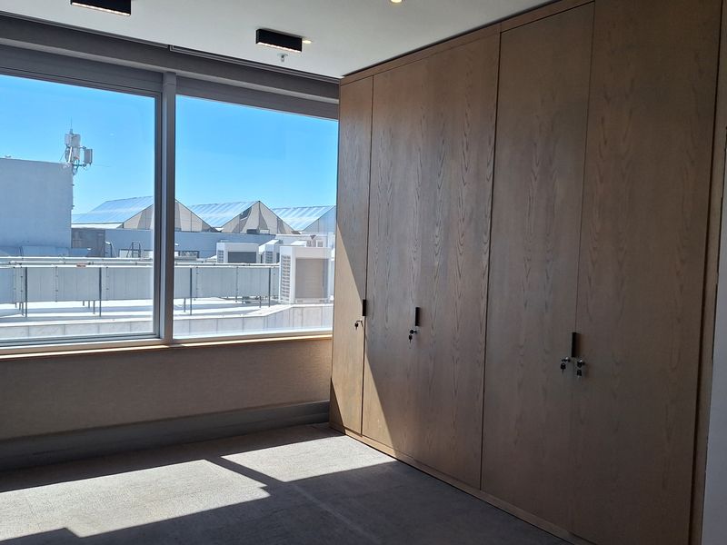 A bright, empty office space featuring extensive built-in wooden storage cabinetry, large windows with natural light, and modern recessed ceiling lighting.