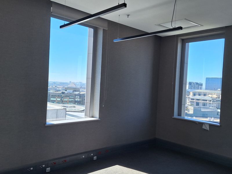 A bright, empty office space featuring modern suspended linear lighting, carpeted floors, and large windows offering expansive city views.