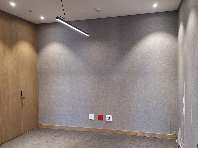 A modern, empty office space featuring fabric-covered walls, built-in wooden cabinetry, carpet tiles, and contemporary recessed and pendant lighting.