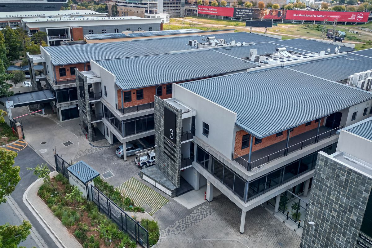 Aerial view of a modern multi-story office complex featuring brick and plaster finishes, metal roofing, and paved parking areas.