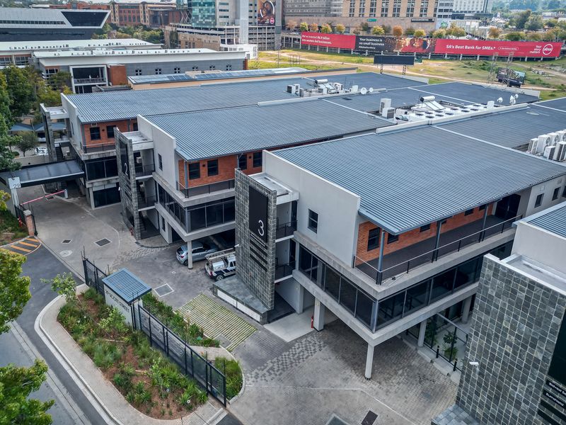 Aerial view of a modern multi-story office complex featuring brick and plaster finishes, metal roofing, and paved parking areas.
