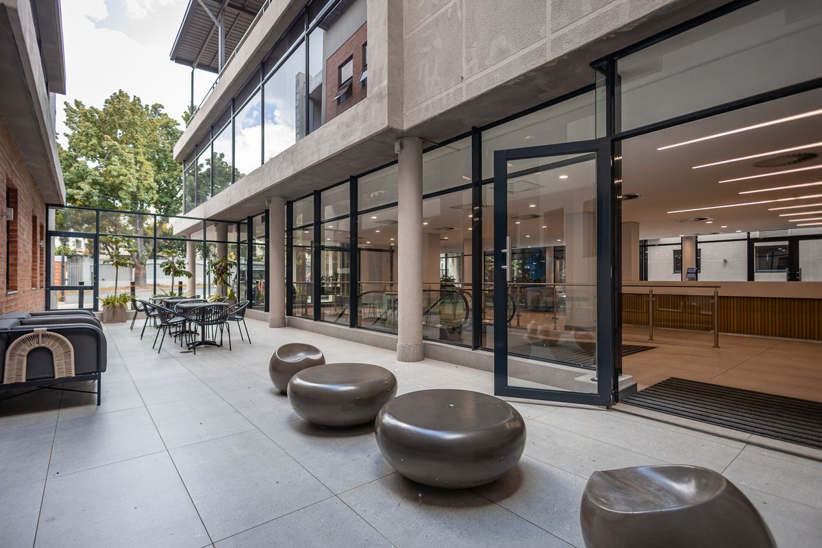A modern outdoor patio area featuring large glass office facades, tiled flooring, and structural concrete columns, providing a seamless transition to the interior workspace.