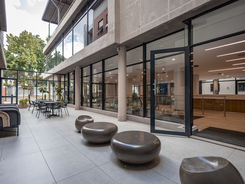 A modern outdoor patio area featuring large glass office facades, tiled flooring, and structural concrete columns, providing a seamless transition to the interior workspace.
