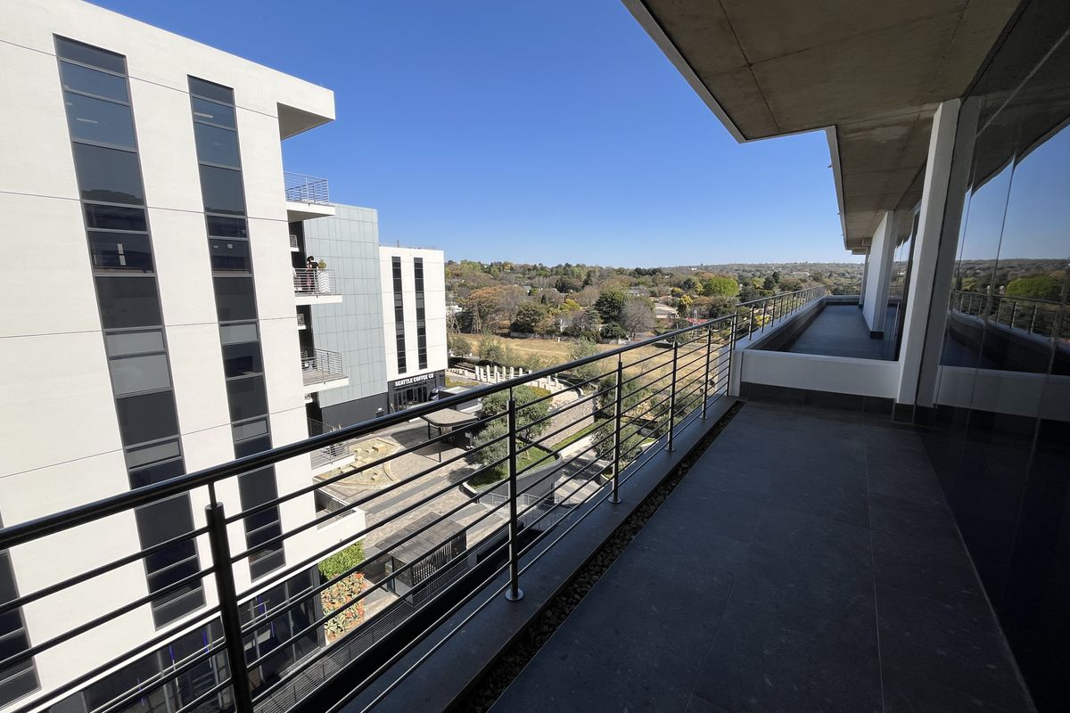 A spacious modern balcony featuring tiled flooring, a sleek stainless steel balustrade, and expansive views of the surrounding office park and landscape.