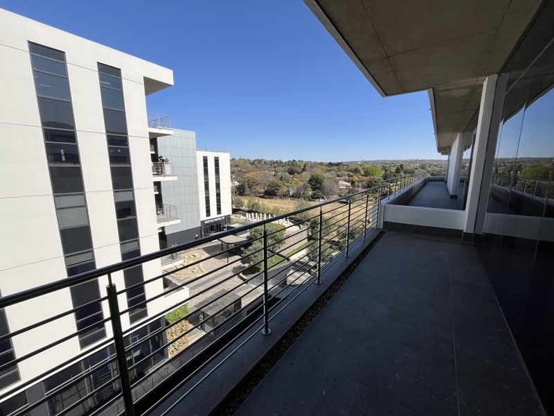 A spacious modern balcony featuring tiled flooring, a sleek stainless steel balustrade, and expansive views of the surrounding office park and landscape.