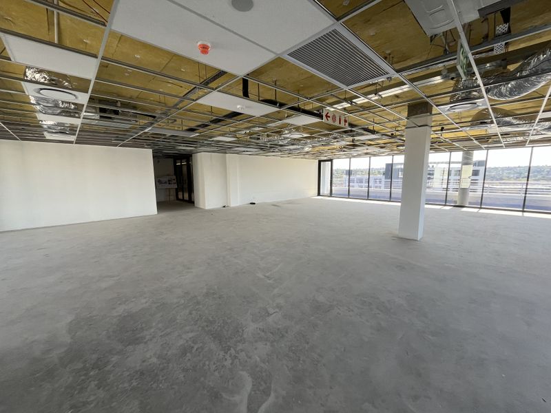 An expansive, open-plan office space in shell condition, featuring floor-to-ceiling windows, exposed ceiling infrastructure, and polished concrete flooring.