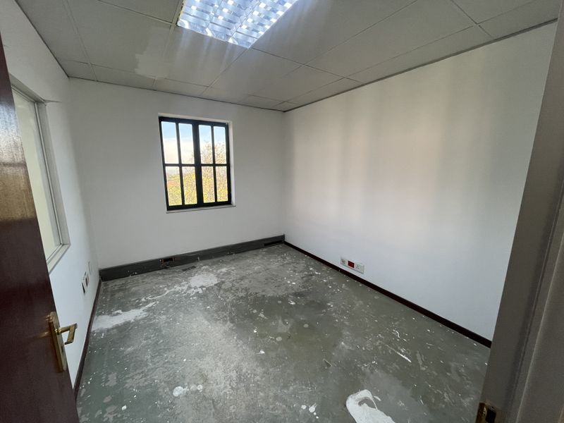 An empty office space featuring a suspended ceiling, recessed lighting, and a window, currently requiring flooring and cosmetic updates.