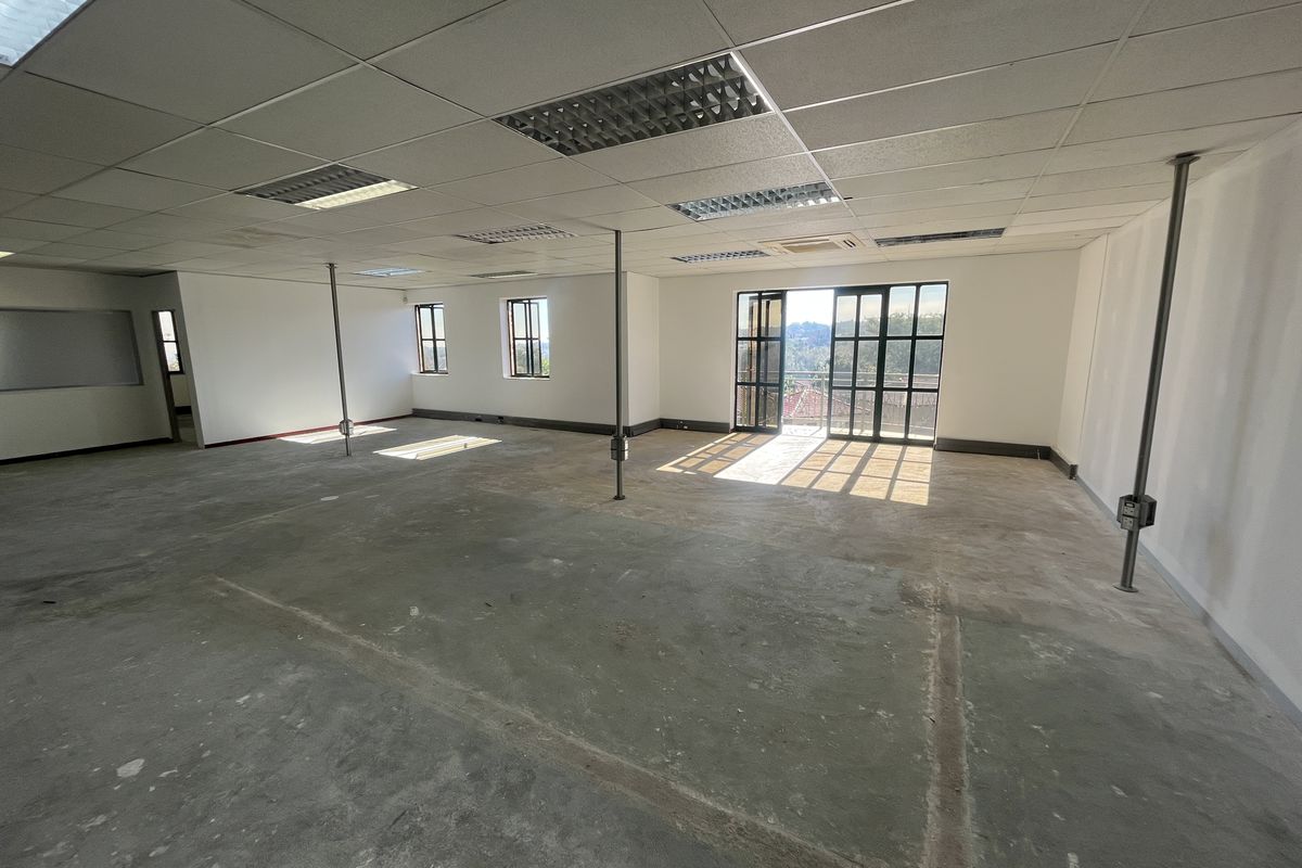 Spacious open-plan office area featuring a suspended ceiling, recessed lighting, metal support columns, and large windows providing natural light and exterior views.