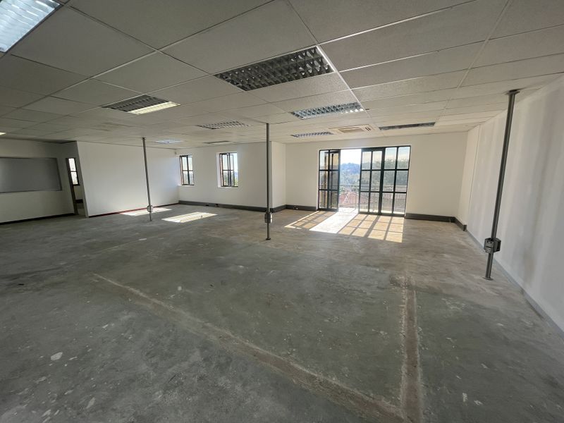 Spacious open-plan office area featuring a suspended ceiling, recessed lighting, metal support columns, and large windows providing natural light and exterior views.