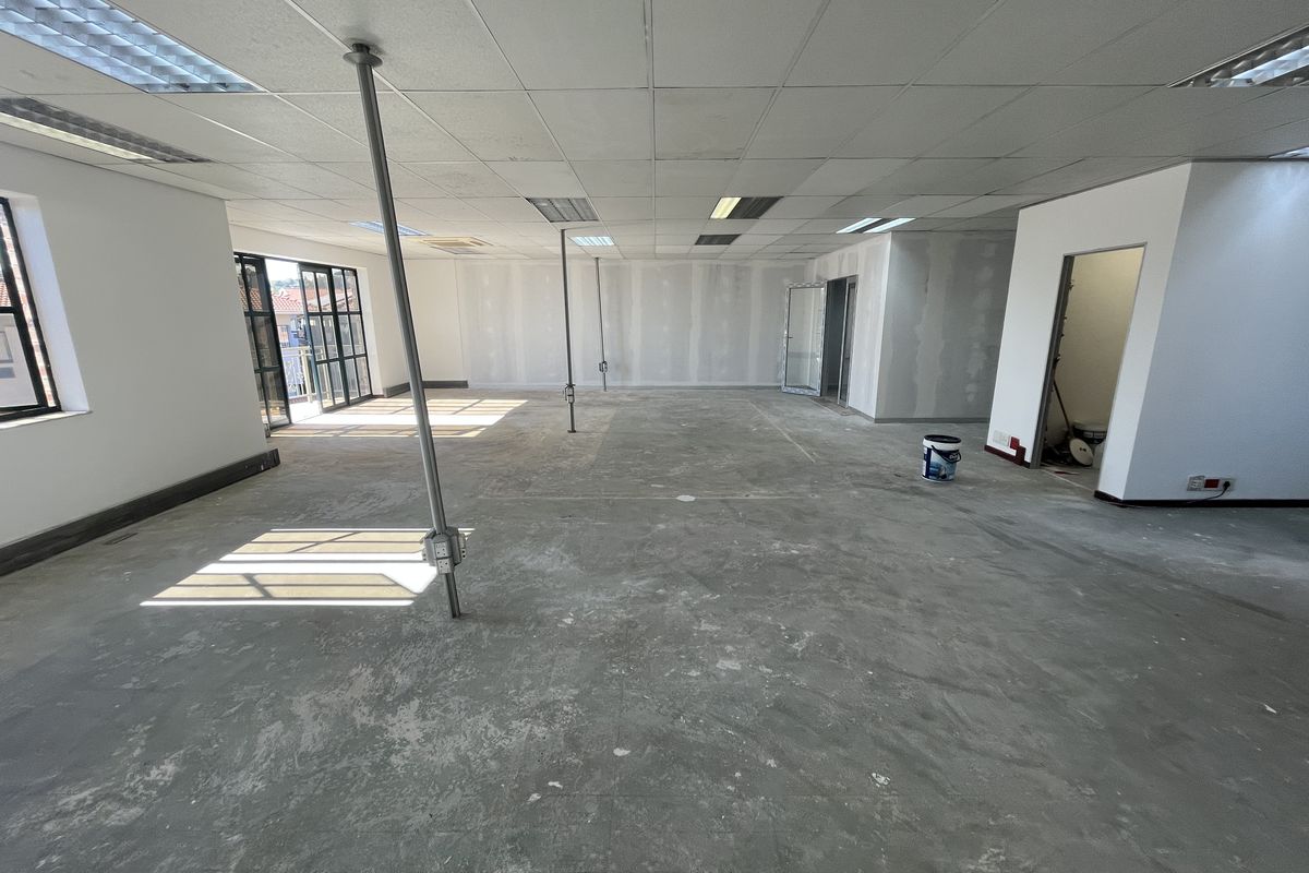 An open-plan office space currently undergoing renovation, featuring a suspended ceiling, concrete flooring, and access to an outdoor balcony area.