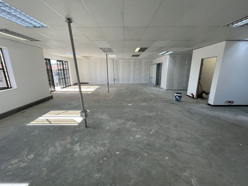 An open-plan office space currently undergoing renovation, featuring a suspended ceiling, concrete flooring, and access to an outdoor balcony area.