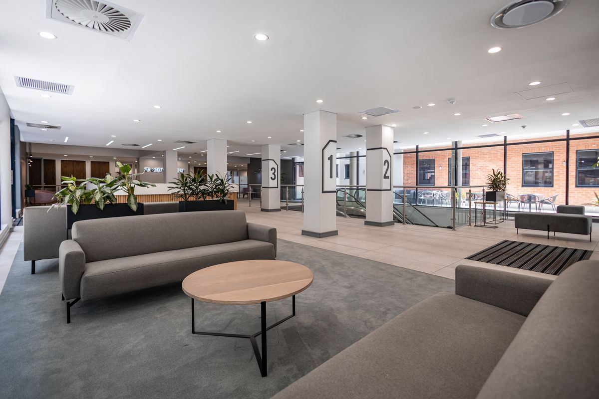 Modern open-plan office lobby featuring high ceilings, recessed lighting, tiled and carpeted flooring, and large glass windows overlooking an exterior courtyard.
