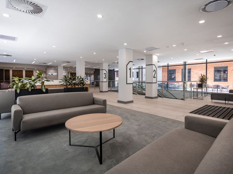 Modern open-plan office lobby featuring high ceilings, recessed lighting, tiled and carpeted flooring, and large glass windows overlooking an exterior courtyard.