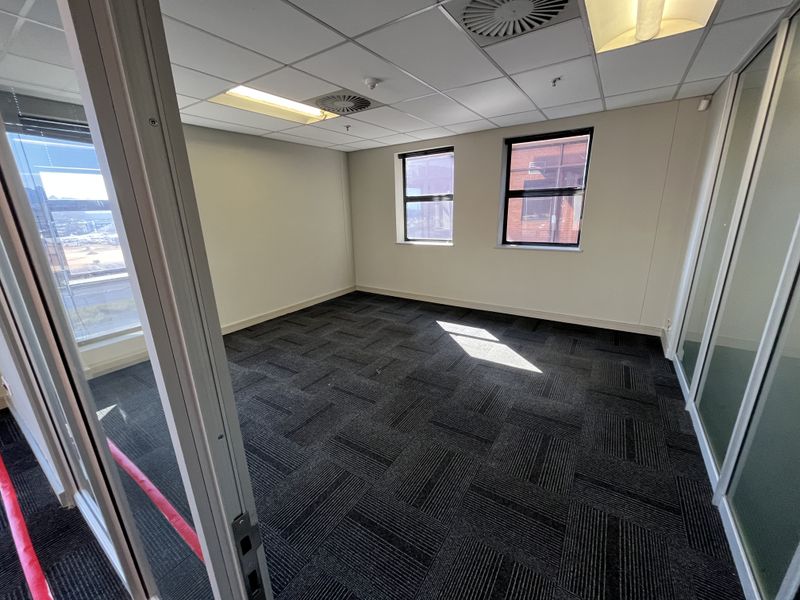 A bright, vacant office space featuring modern carpet tiles, glass partition walls, and a suspended ceiling with integrated lighting and air conditioning.