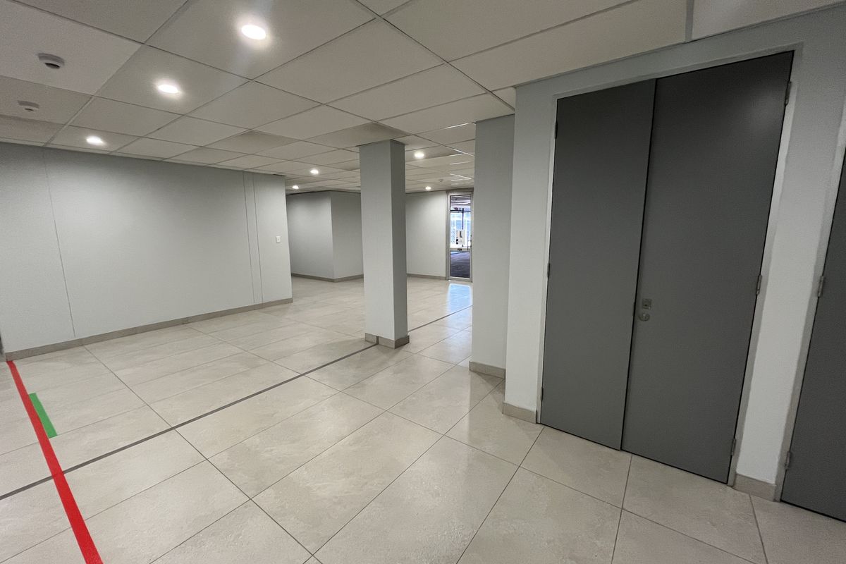 A spacious, empty office area featuring neutral ceramic floor tiles, a modern suspended ceiling with recessed lighting, and clean, minimalist wall finishes.