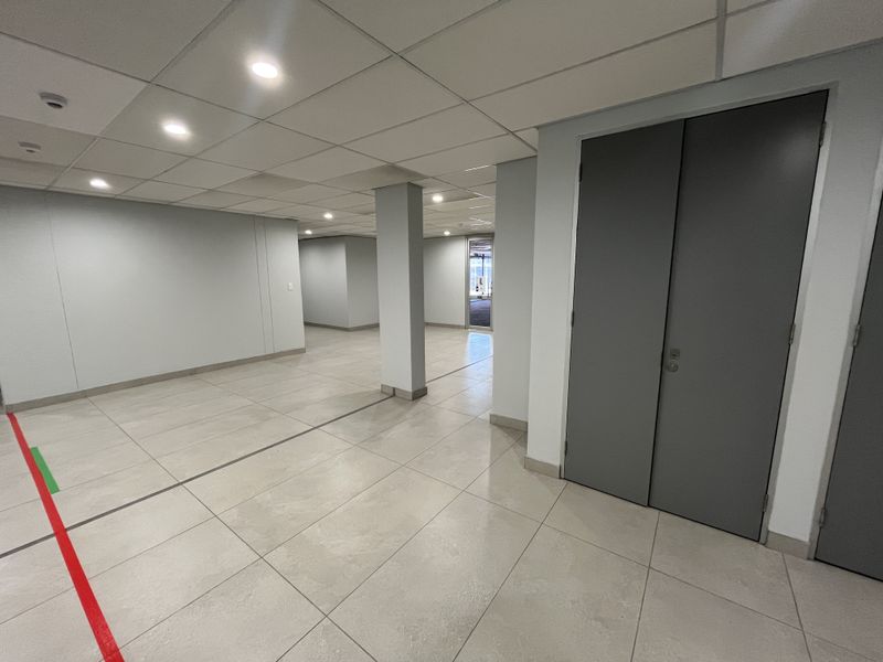 A spacious, empty office area featuring neutral ceramic floor tiles, a modern suspended ceiling with recessed lighting, and clean, minimalist wall finishes.