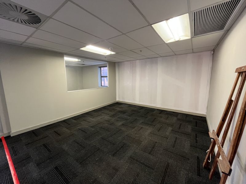 An empty office space featuring carpet tiles, a suspended ceiling with recessed lighting, and an internal window, currently undergoing wall finishing.