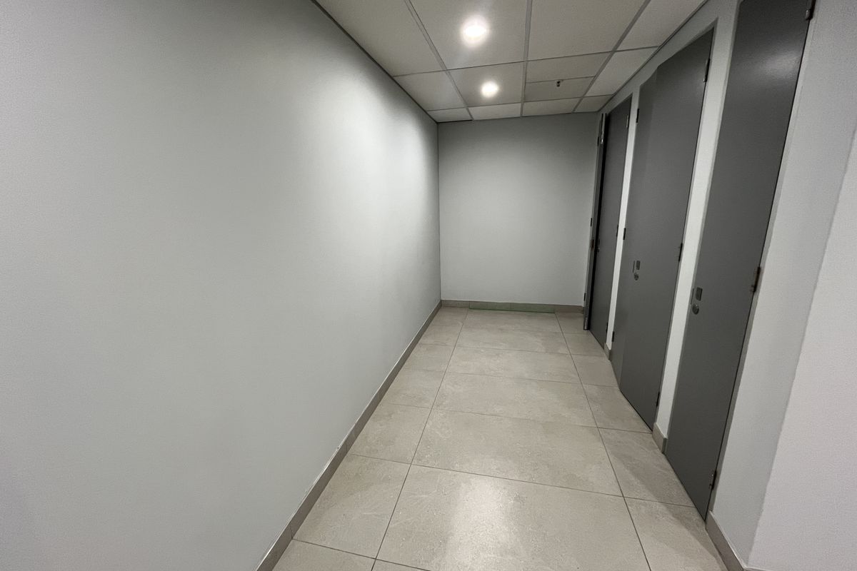 A clean, modern office hallway featuring neutral grey walls, large format floor tiles, and a suspended ceiling with integrated lighting.