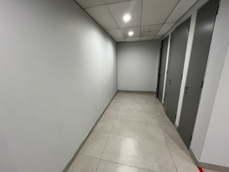 A clean, modern office hallway featuring neutral grey walls, large format floor tiles, and a suspended ceiling with integrated lighting.