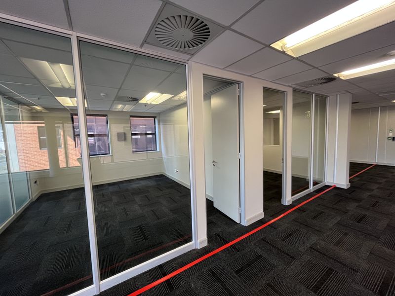 Modern office space featuring glass partitioning, suspended ceilings with recessed lighting, and dark carpet tiles in a clean, professional environment.