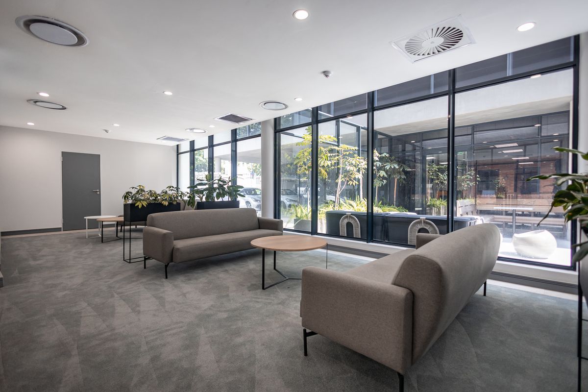 Modern office lounge area featuring floor-to-ceiling glass windows, recessed lighting, and integrated ceiling air conditioning units with a view of an outdoor courtyard.
