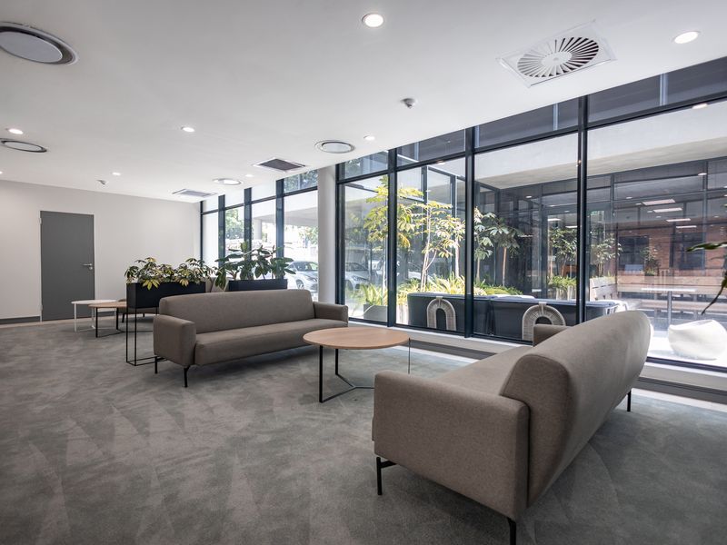 Modern office lounge area featuring floor-to-ceiling glass windows, recessed lighting, and integrated ceiling air conditioning units with a view of an outdoor courtyard.