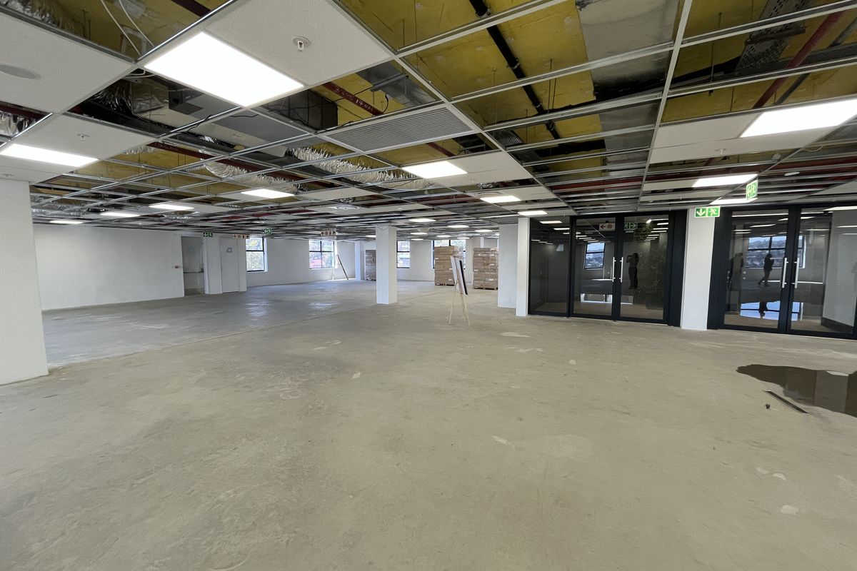 Open-plan office space currently under renovation, featuring exposed ceiling services, concrete flooring, and glass-partitioned meeting rooms.