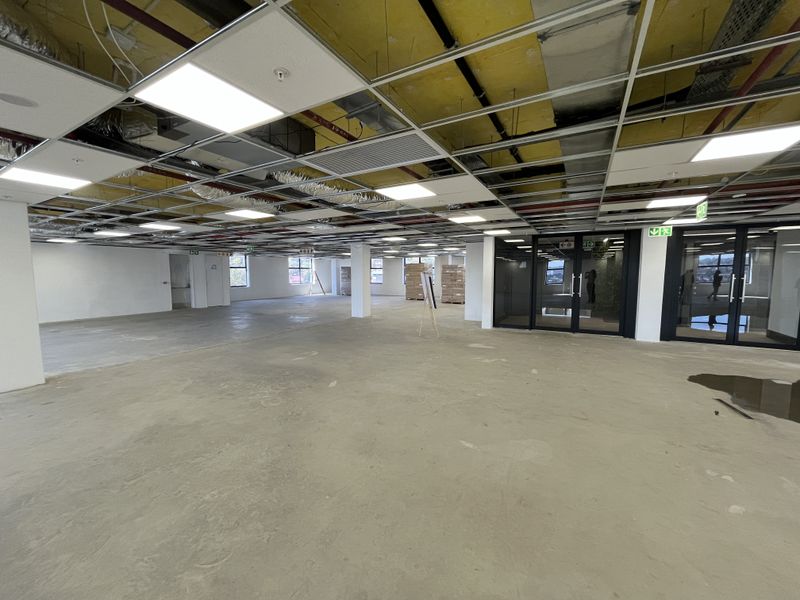 Open-plan office space currently under renovation, featuring exposed ceiling services, concrete flooring, and glass-partitioned meeting rooms.