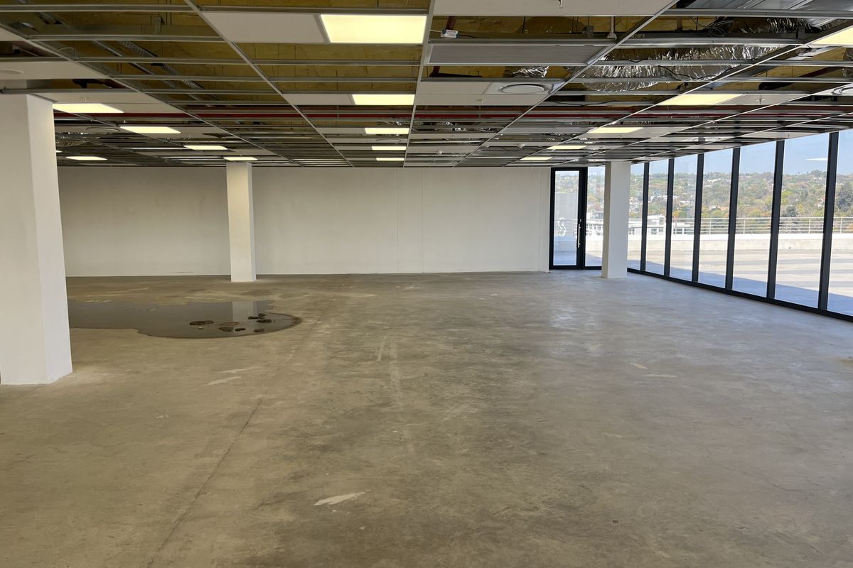 An expansive, open-plan office space featuring floor-to-ceiling windows, concrete flooring, and an unfinished suspended ceiling grid requiring completion.