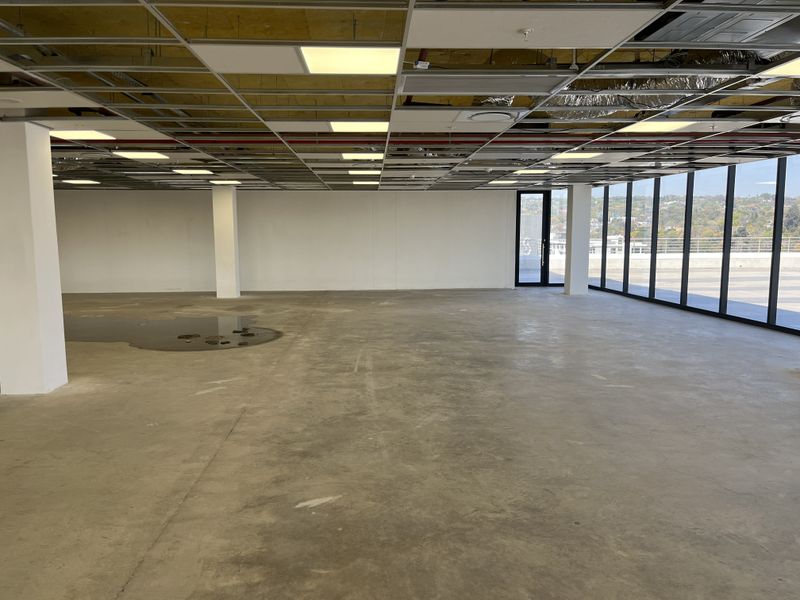 An expansive, open-plan office space featuring floor-to-ceiling windows, concrete flooring, and an unfinished suspended ceiling grid requiring completion.