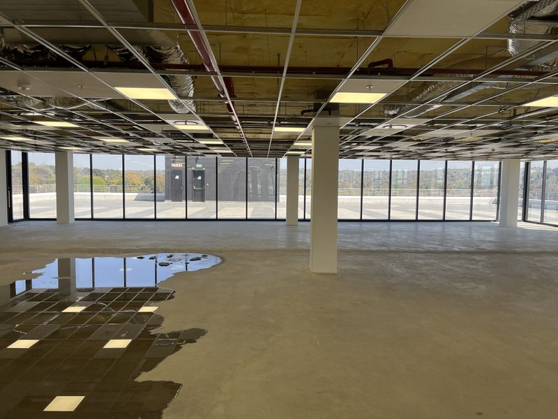 Spacious open-plan office area featuring floor-to-ceiling windows, exposed ceiling infrastructure, and structural columns, currently in an unfinished state.