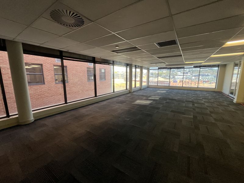 A spacious, empty open-plan office area featuring large windows, carpeted flooring, and a suspended ceiling with integrated lighting.