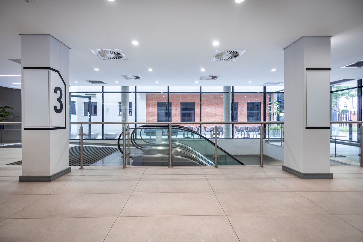 Modern office building lobby area featuring tiled flooring, a central escalator, and large glass windows overlooking an outdoor courtyard.