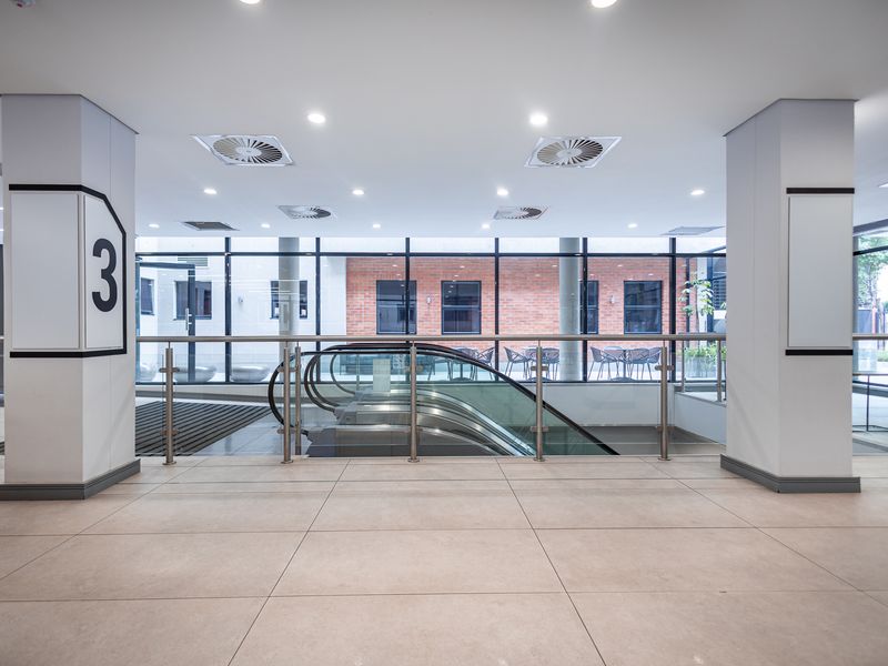 Modern office building lobby area featuring tiled flooring, a central escalator, and large glass windows overlooking an outdoor courtyard.
