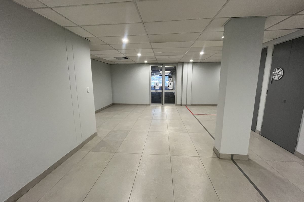 A spacious, vacant office area featuring neutral ceramic floor tiles, a suspended ceiling with recessed lighting, and a glass entrance door.