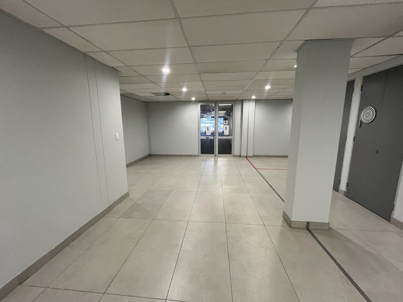 A spacious, vacant office area featuring neutral ceramic floor tiles, a suspended ceiling with recessed lighting, and a glass entrance door.