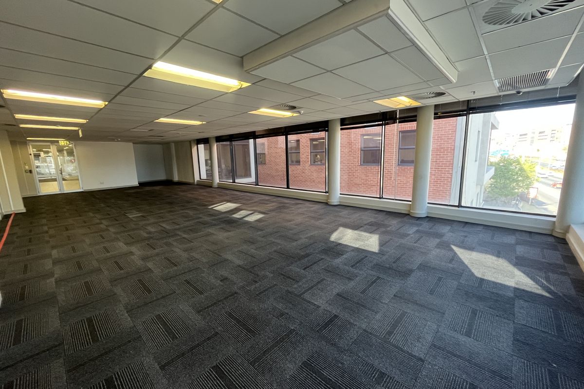 A spacious, open-plan office area featuring carpet tiles, a suspended ceiling with recessed lighting, and large windows providing ample natural light.