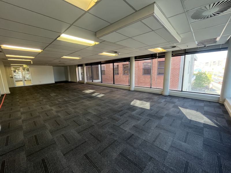 A spacious, open-plan office area featuring carpet tiles, a suspended ceiling with recessed lighting, and large windows providing ample natural light.