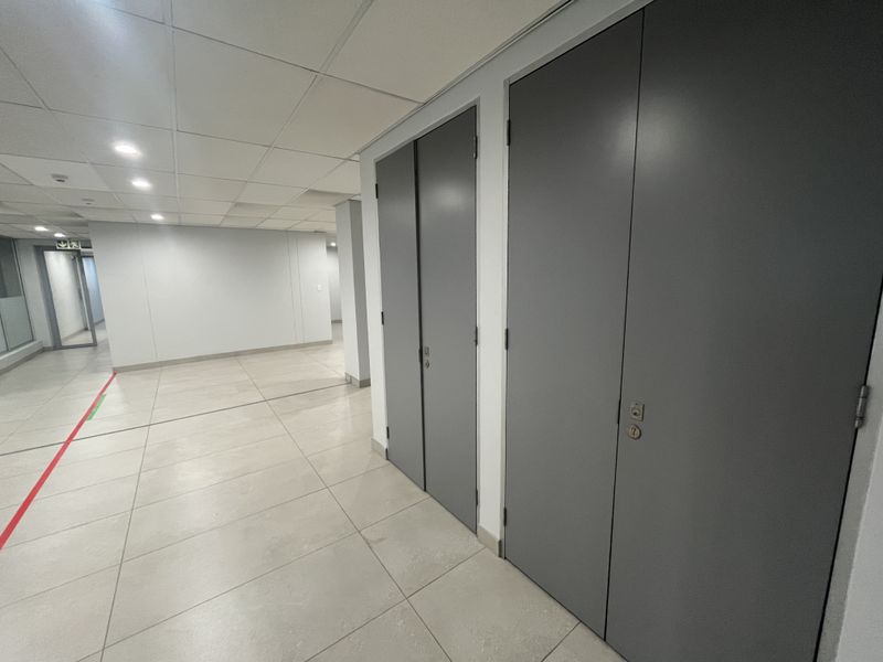 A clean, modern office hallway featuring neutral tiled flooring, a suspended ceiling with recessed lighting, and sleek full-height grey doors.