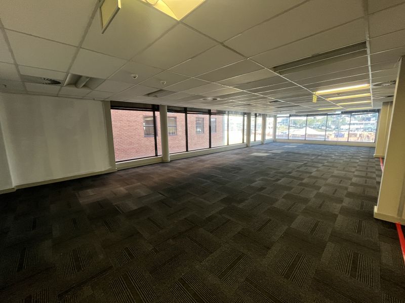 A spacious, vacant open-plan office area featuring large windows, suspended ceilings with recessed lighting, and durable carpet tile flooring.