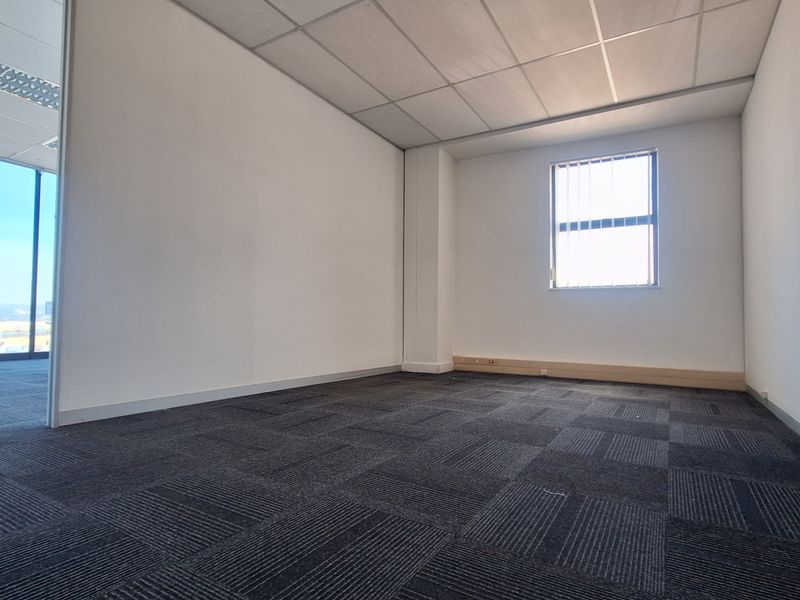 A vacant, well-lit office space featuring neutral walls, dark carpet tiles, and a suspended ceiling with a window providing natural light.