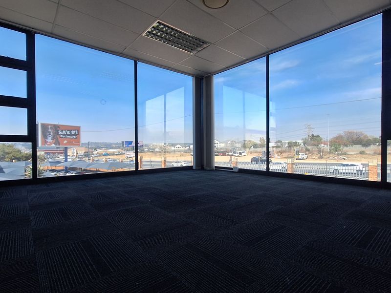 A bright, empty office space featuring large floor-to-ceiling windows with expansive views, carpeted flooring, and a suspended ceiling with recessed lighting.