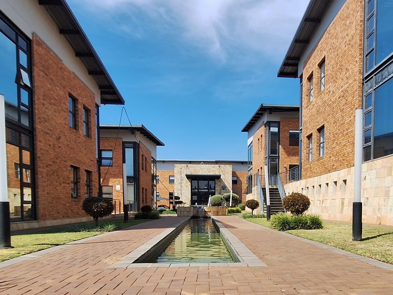 A well-maintained office complex courtyard featuring brick buildings, large glass windows, a central water feature, and paved walkways.