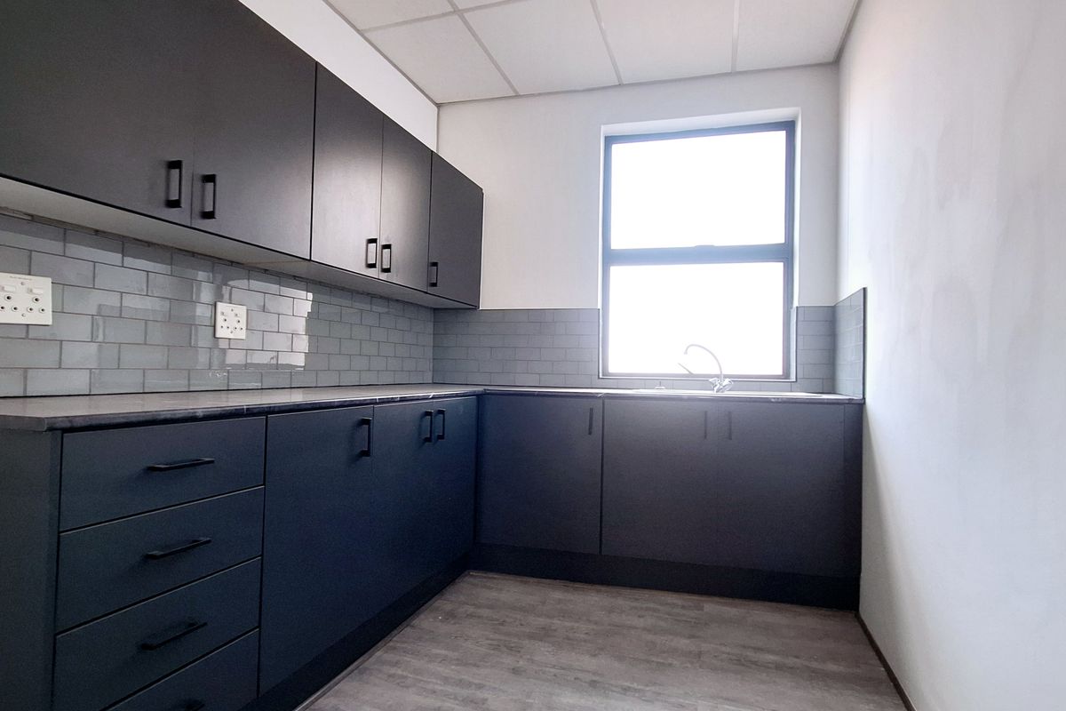 A modern office kitchenette featuring dark cabinetry, subway tile backsplash, laminate countertops, and a stainless steel sink in a clean, empty space.