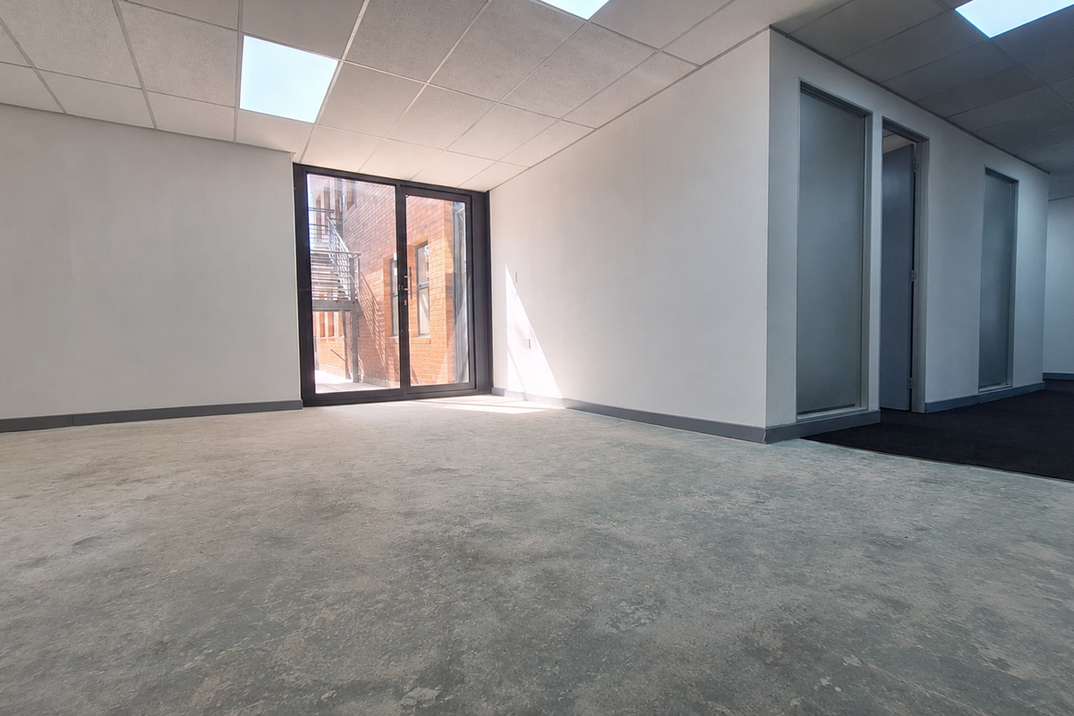 An empty, open-plan office space featuring a suspended ceiling, recessed lighting, and glass partitions, with direct access to an external staircase.