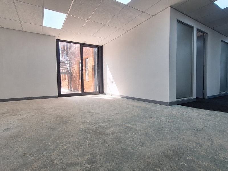 An empty, open-plan office space featuring a suspended ceiling, recessed lighting, and glass partitions, with direct access to an external staircase.