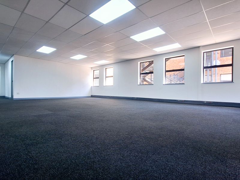 A spacious, open-plan office area featuring neutral carpet tiles, a suspended ceiling with recessed lighting, and large windows providing ample natural light.