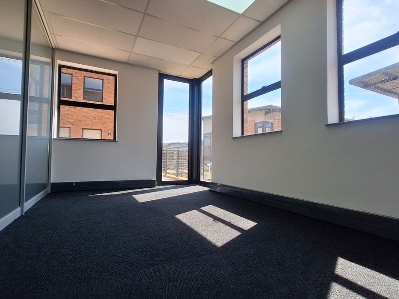 A bright, empty office space featuring large windows, carpeted flooring, and a modern glass partition wall, offering a clean and professional work environment.
