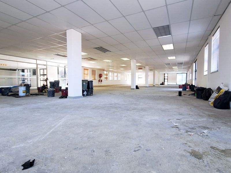 A large, open-plan office space featuring a suspended ceiling, recessed lighting, and structural columns, currently in a shell condition awaiting final flooring installation.
