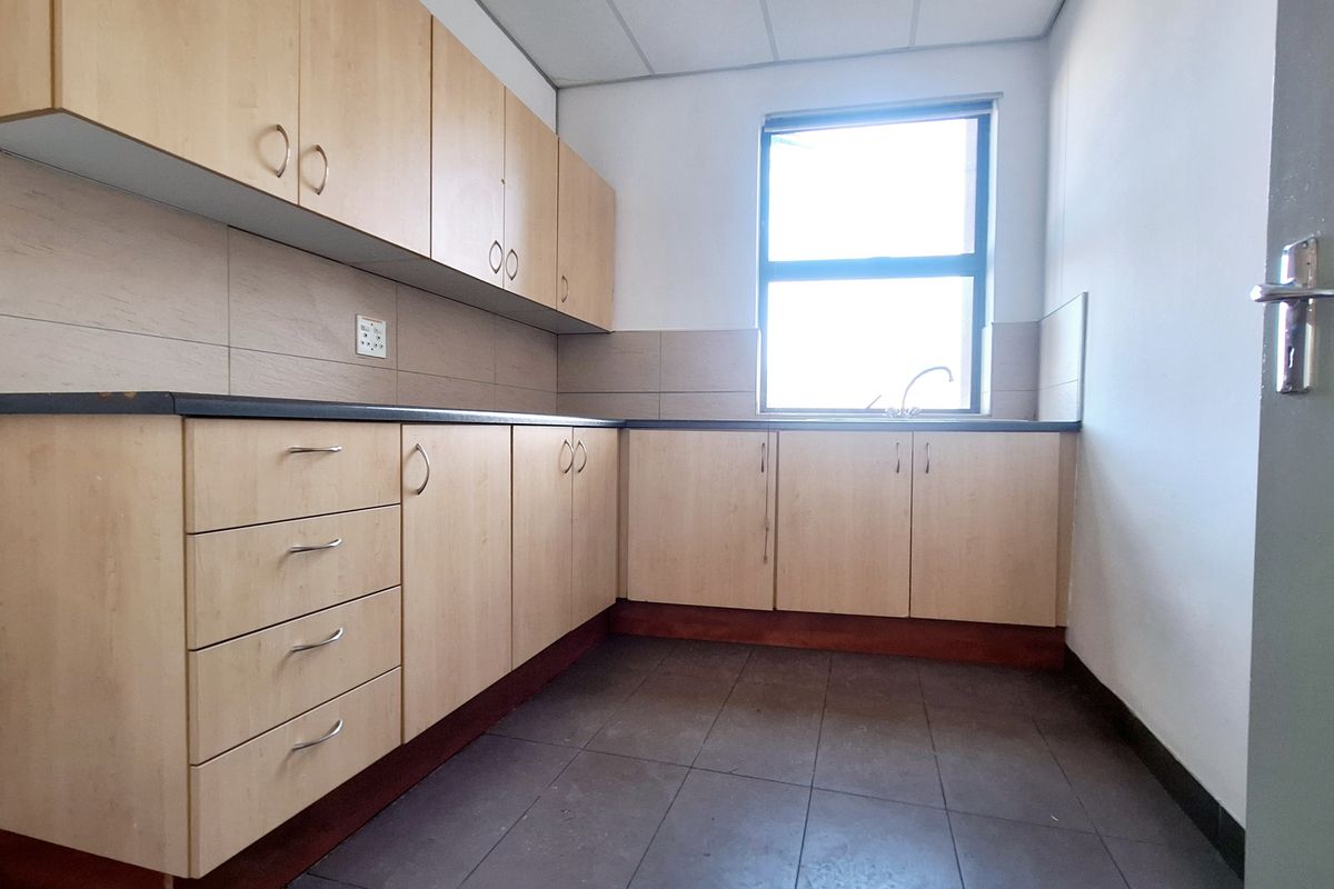 A functional office kitchenette featuring light wood-finish cabinetry, dark laminate countertops, a stainless steel sink, and tiled flooring.